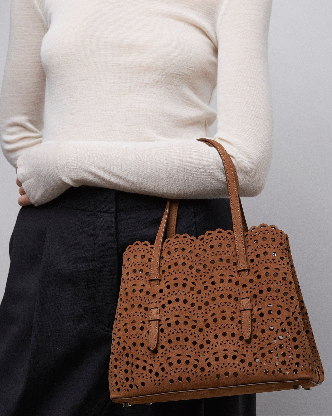 Person wearing a white sweater and holding a brown perforated handbag against a plain background