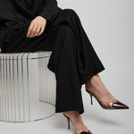 Person wearing a black outfit with high-heeled shoes sitting on a wire stool against a white background
