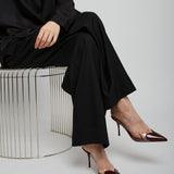 Person wearing a black outfit with high-heeled shoes sitting on a wire stool against a white background