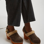 Brown suede clogs with fur trim worn with black jeans on a white background