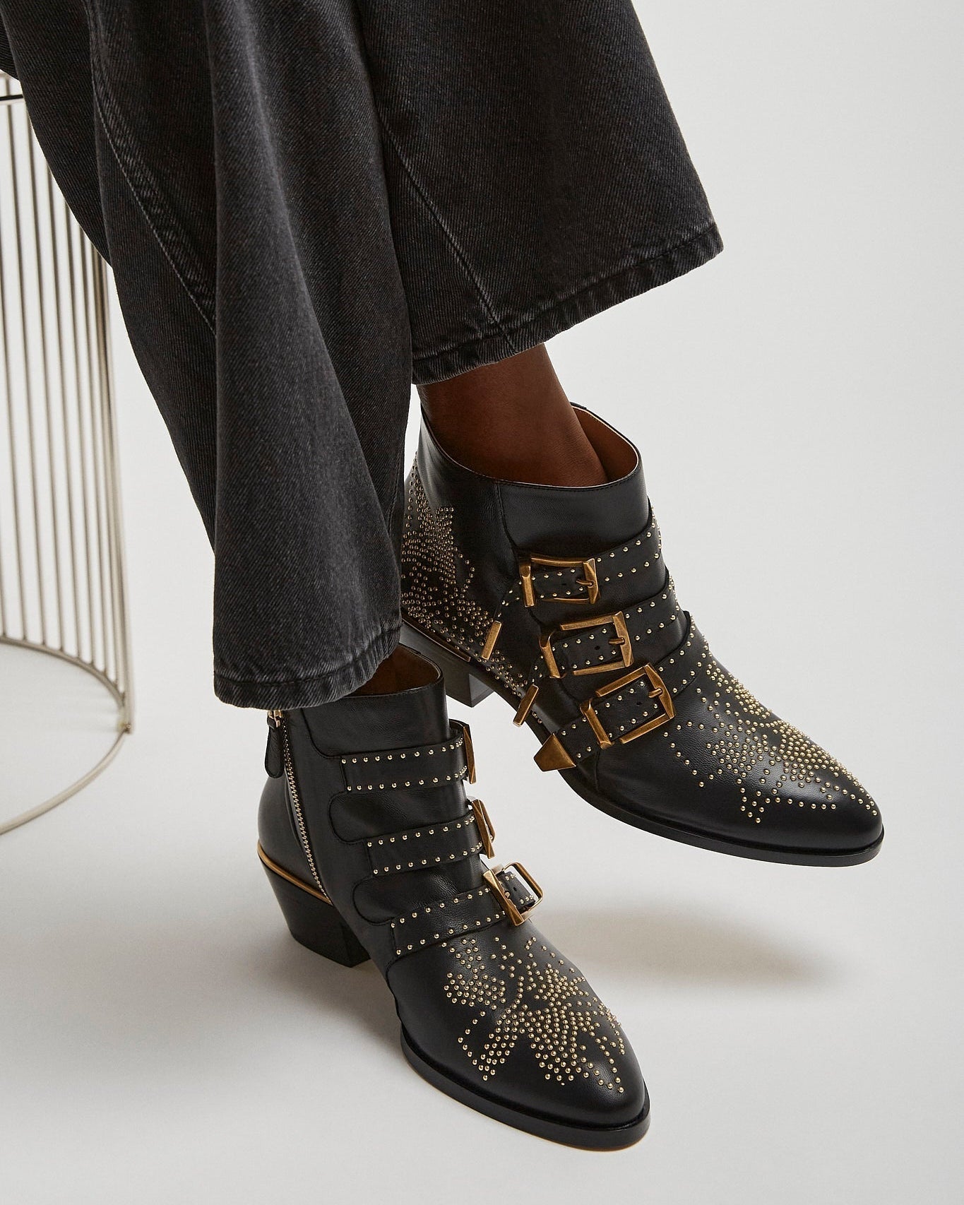 Black studded boots with gold buckles worn by a person sitting on a white chair.