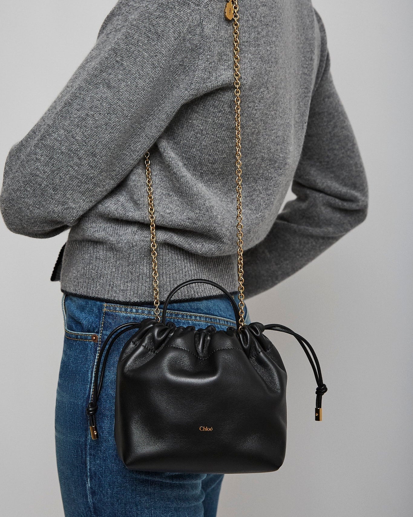 Person wearing a gray sweater with a black handbag featuring a gold chain strap on a plain background