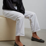 Person wearing a black shirt, white pants, and black studded flats sitting on a beige block.