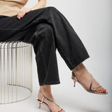 Person wearing black pants and transparent high-heeled sandals sitting on a wire stool against a white background.