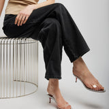 Person wearing a beige top and black pants sitting on a wire stool against a plain background