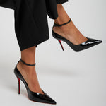 Black high-heeled shoes with red soles worn by a person in black pants on a light gray background