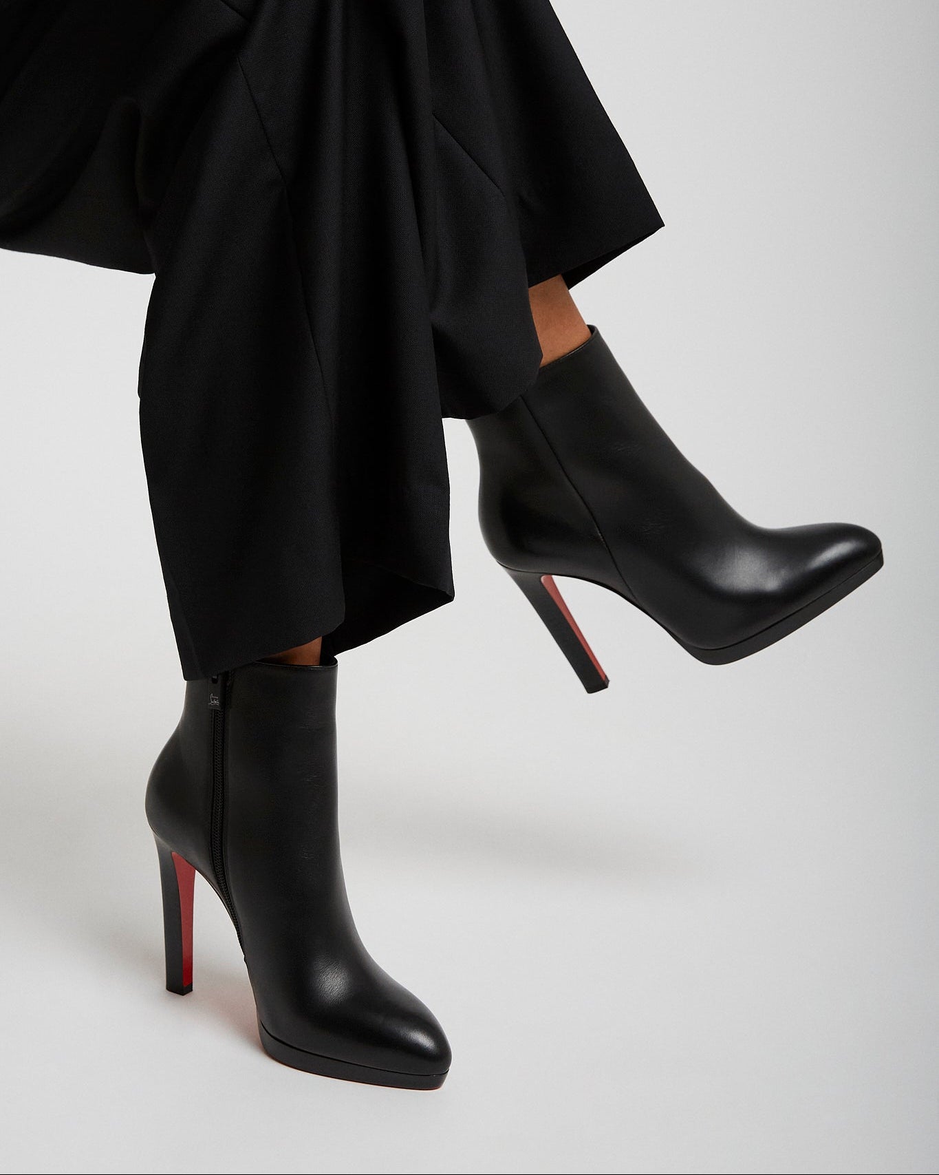 Black high-heeled boots with red soles worn by a person sitting on a light gray background