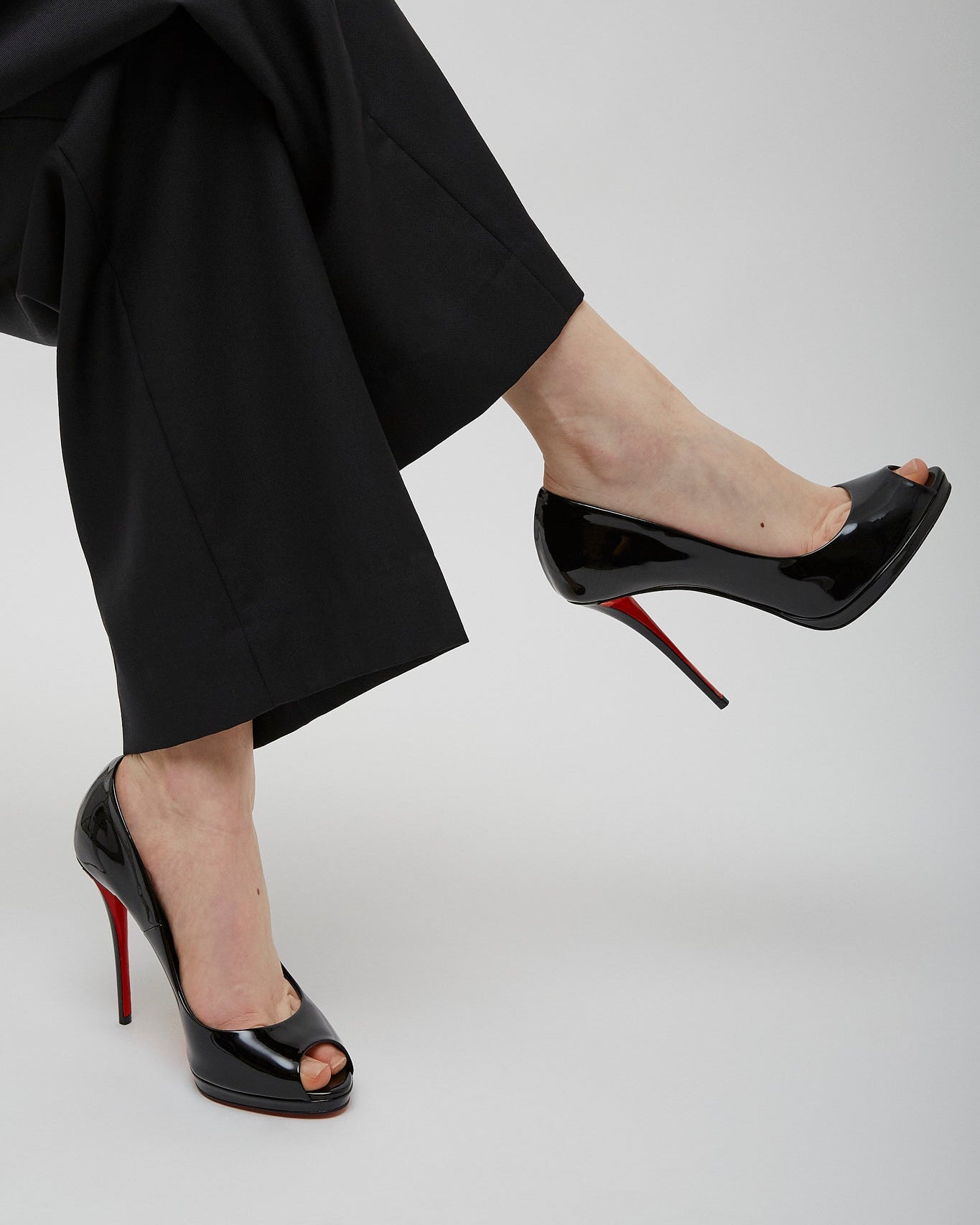 Black high-heeled shoes with red soles worn by a person in black pants on a light gray background