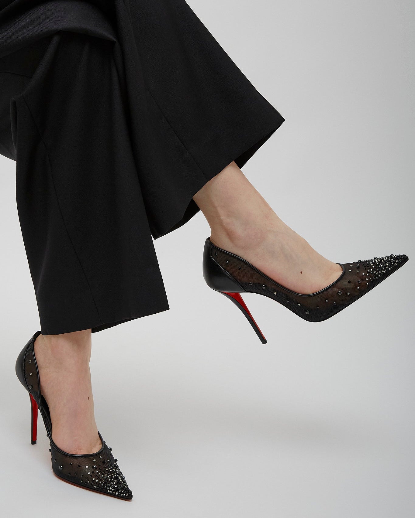Black high-heeled shoes with red soles worn by a person in black pants on a light gray background