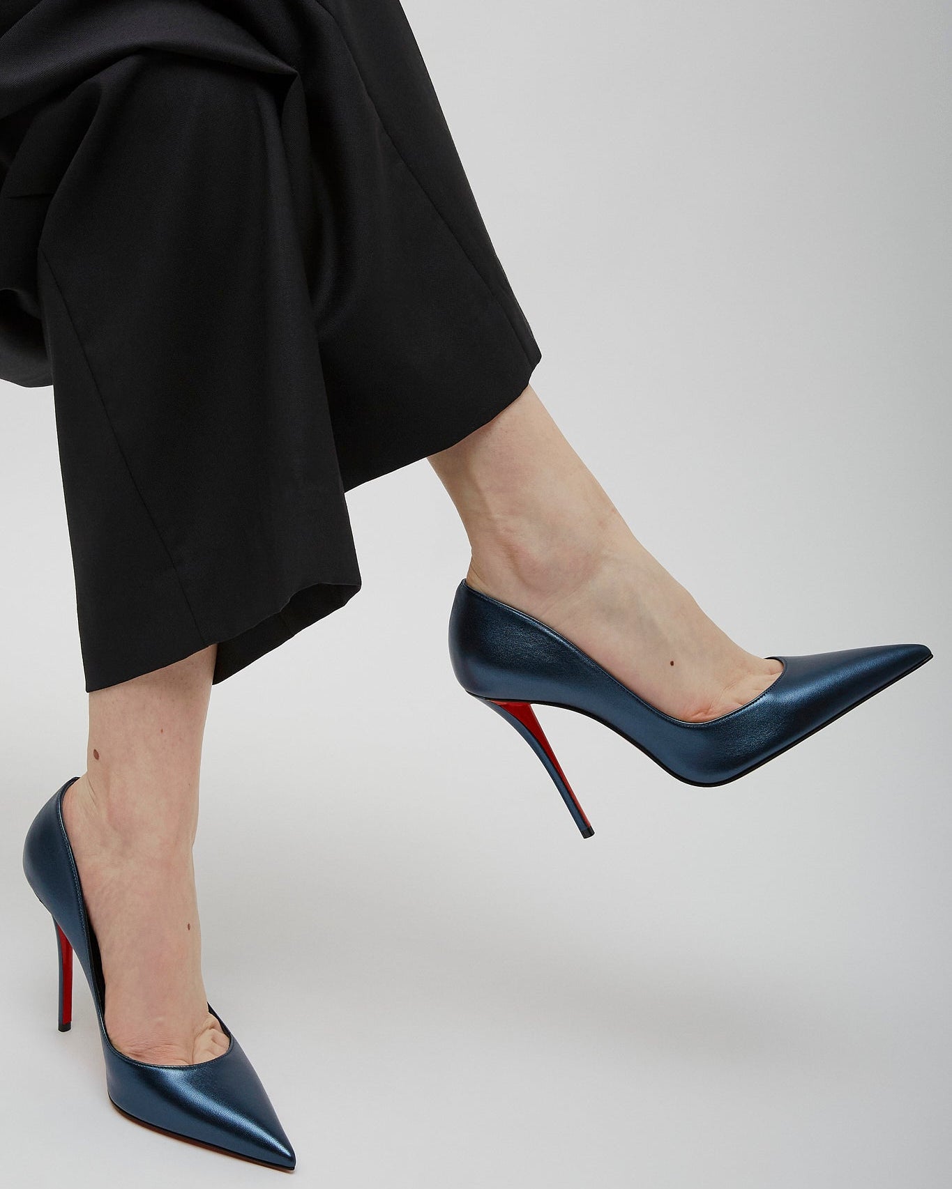 Navy high-heeled shoes with red soles worn by a person in black pants on a light gray background