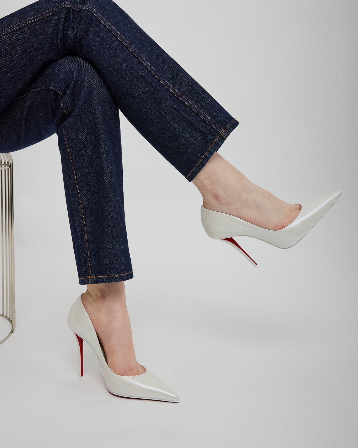 Person wearing dark jeans and white high-heeled shoes with red soles on a light gray background