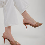 Gold high-heeled shoes with red soles worn by a person in white pants on a light gray background