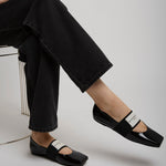 Person wearing black patent leather shoes with a white brand logo, sitting on a chair.