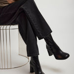 Person wearing black boots and black pants sitting on a wire stool against a white background