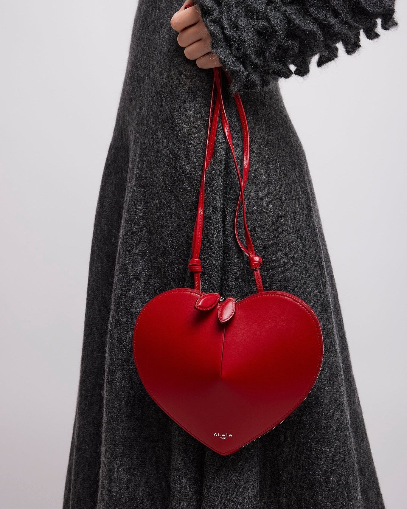 Red heart-shaped handbag held by a person wearing a gray dress against a white background