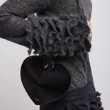 Person wearing a dark gray outfit with ruffled sleeves and holding a black heart-shaped clutch.