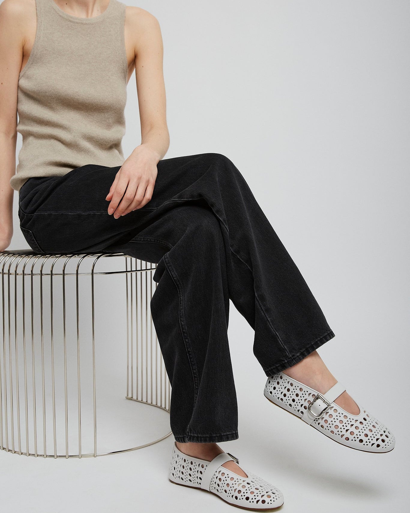 Person wearing a beige sleeveless top and black pants, sitting on a wire stool with white shoes.