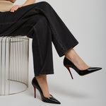 Person wearing black high-heeled shoes with red soles, sitting on a wire stool against a plain background.