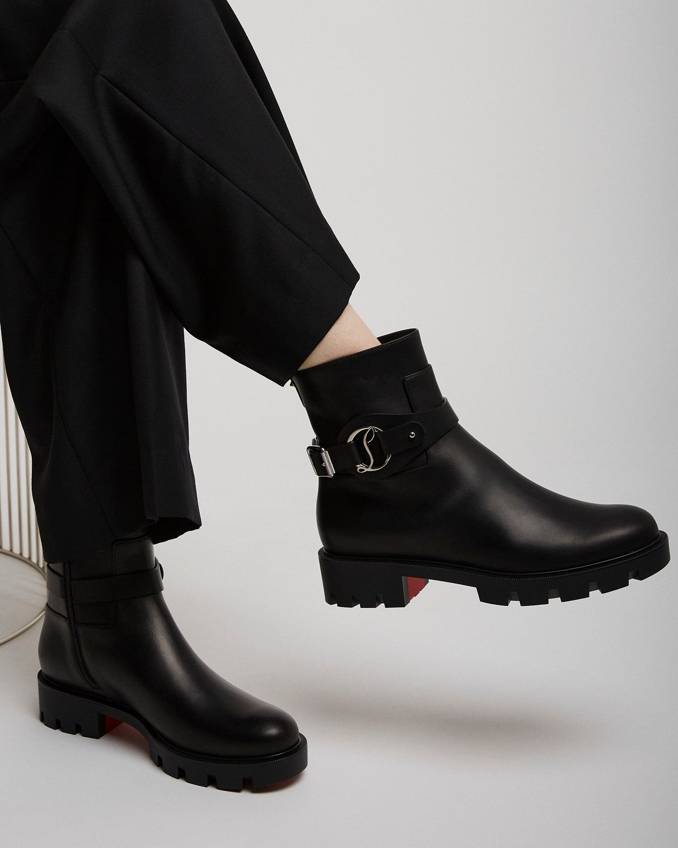 Black leather boots with a brand logo worn by a person sitting on a white chair.